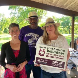 Three people smiling for the camera at a picnic shelter. White woman with cropped brown hair. African American man with white hat. White female with long blonde hair wearing straw sun hat and holding volunteer sign.